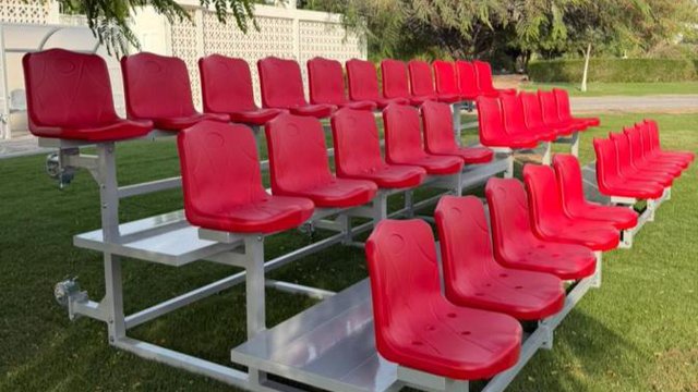 Two-tier aluminium-frame spectator bleachers with red plastic seats, the kind that lines a community football touchline