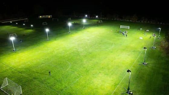A football pitch lit by tall floodlight towers after dark, training continuing under the lights well after sunset